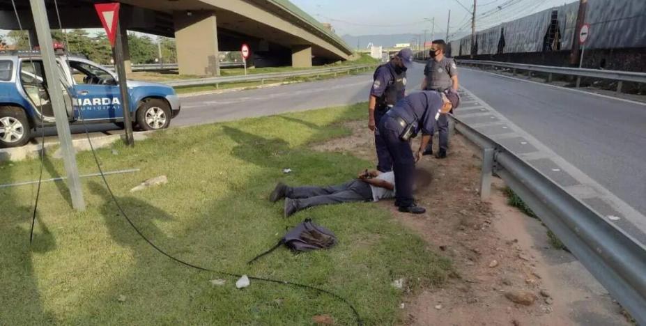 Viatura da guarda municipal parada à esquerda. Guarda e policiais estão em pé ao lado de homem deitado que está dominado. #paratodosverem