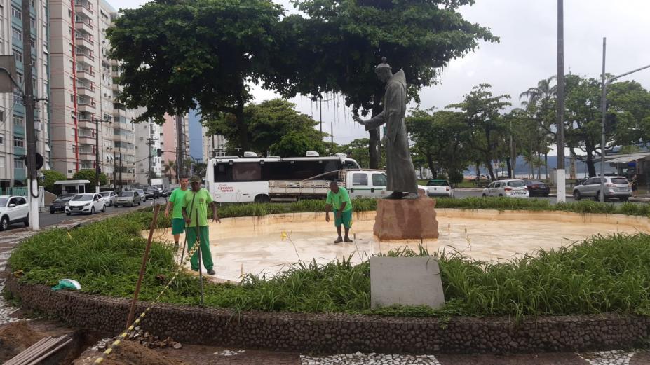 três operários uniformizados estão dentro de área da fonte, sem água. A estátua de santo antônio está ao centro. A área da fonte é rodeada por vegetação. #paratodosverem