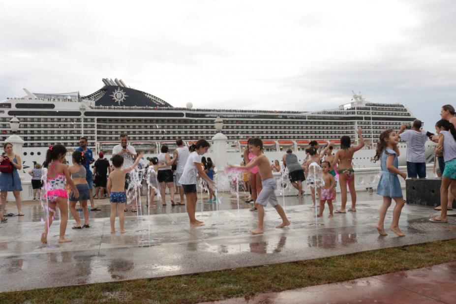 Crianças brincam como água da fonte interativa com navio de cruzeiro passando ao fundo. #pracegover