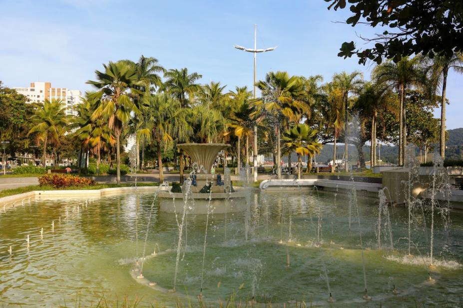 Vista geral de praça com chafariz e palmeiras ao fundo. #paratodosverem 