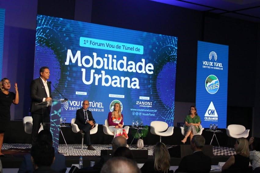 palco com pessoas sentadas e o prefeito em pé falando ao público. Ao fundo, um painel onde se lê Mobilidade Urbana - #paratodosverem
