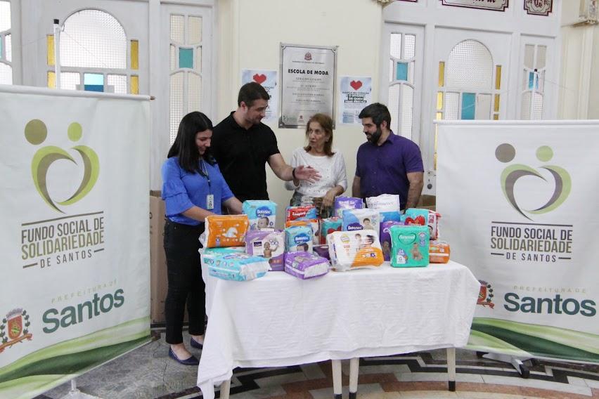 pacotes de fraldas sobre uma mesa e quatro pessoas atrás. Nas laterais, dois banners do fundo social. #paratodosverem