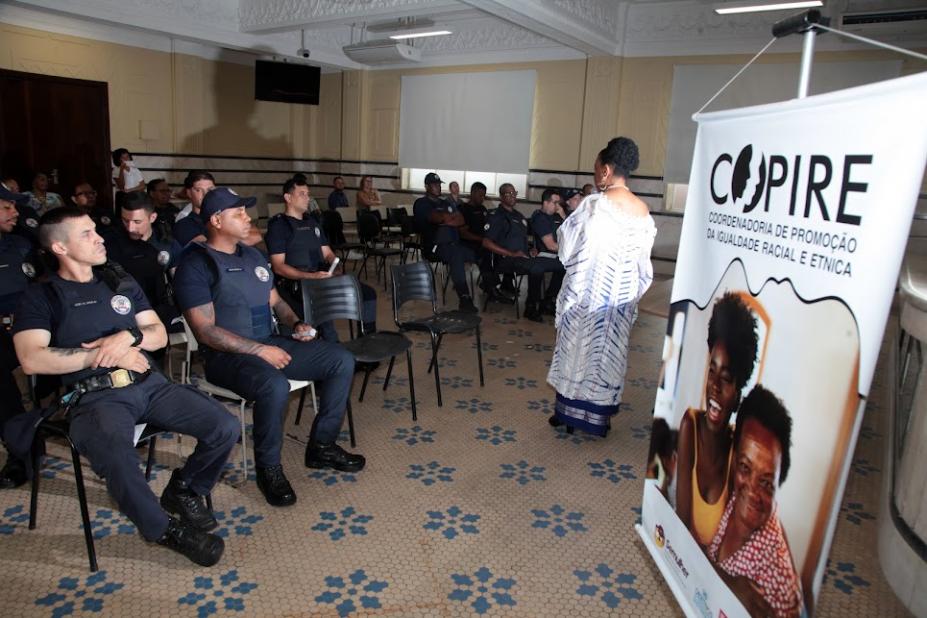 guardas estão sentados ouvindo palestra de uma mulher que está de costas para a foto. À frente, um banner da copire. #paratodosverem 