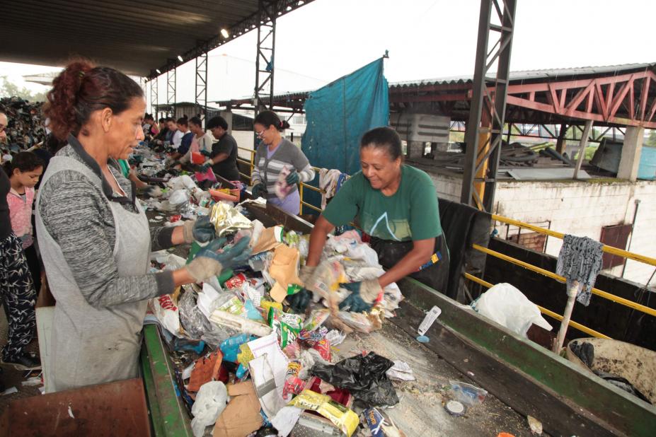 #pracegover Com luvas, duas mulhers separam lixo reciclável sobre esteira em galpão 