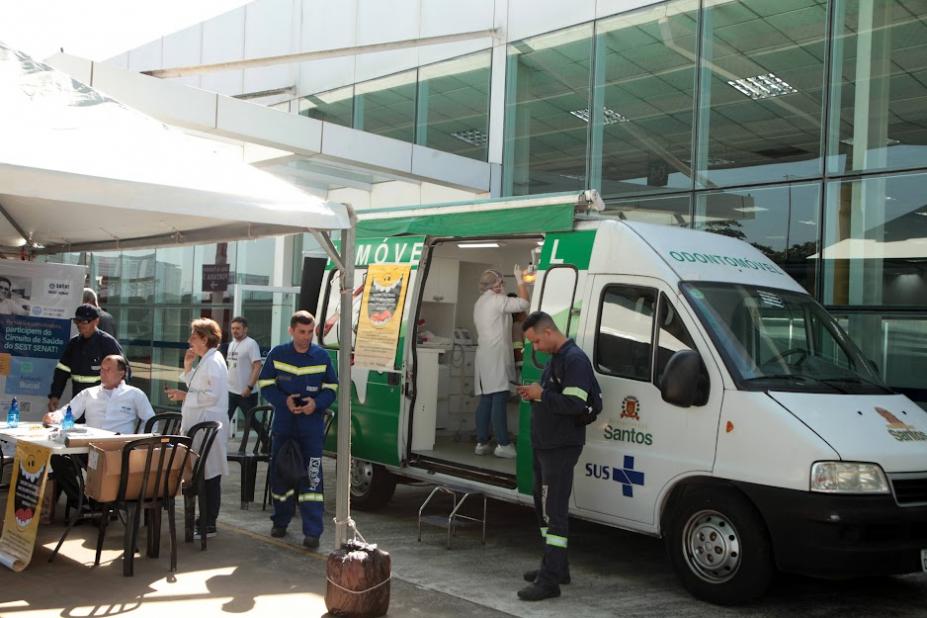 van odontológica está parada em frente a terminal. diante dela há uma tenda com pessoas usando avental branco. Trabalhadores portuários estão uniformizados. #paratodosverem