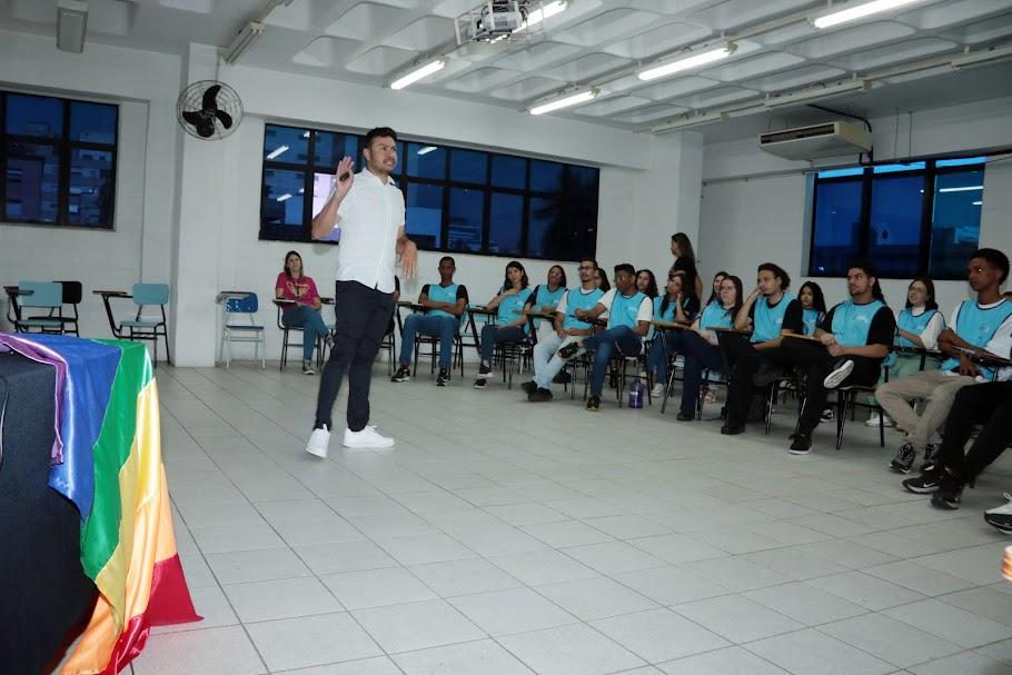 em primeiro plano, mesa com bandeira da causa lgbt+. Ao fundo, homem em pé da palestra para jovens sentados. #paratodosverem 