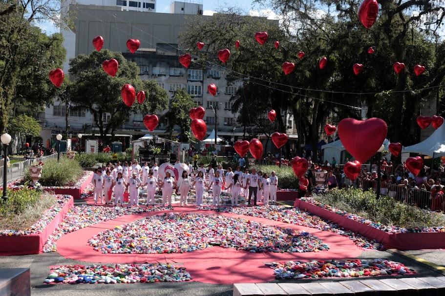milhares de corações de tecido dispostos no chão da praça mauá, com pessoas vestidas de branco e de mãos dadas ao fundo. Em primeiro plano, decoração de corações pendurados em fios. #paratodosverem 