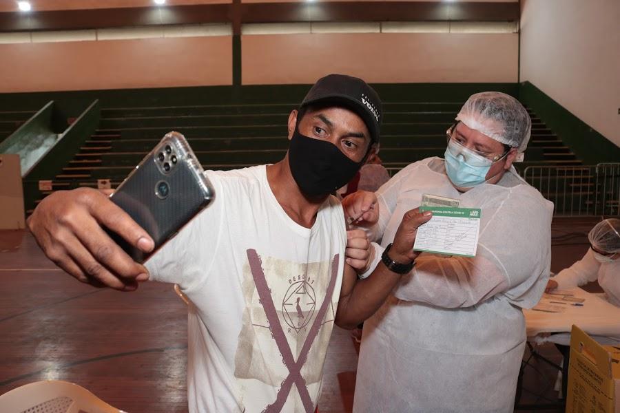 Homem de boné segurando celular recebe aplicação de vacina de enfermeiro. #paratodosverem