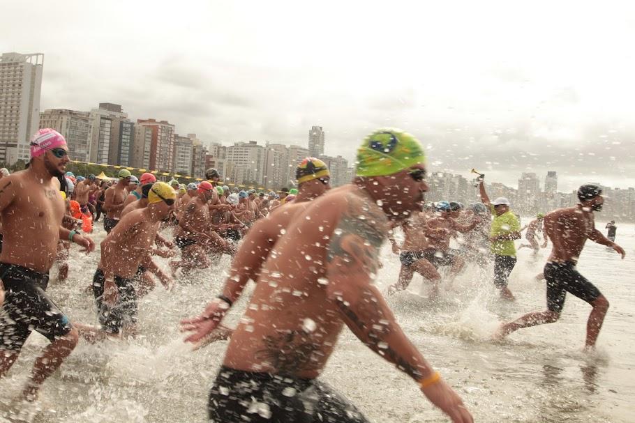 homens entram no mar para prova aquática. Eles estão correndo na beira da água indo em direção ao fundo para nadar. #paratodosverem 
