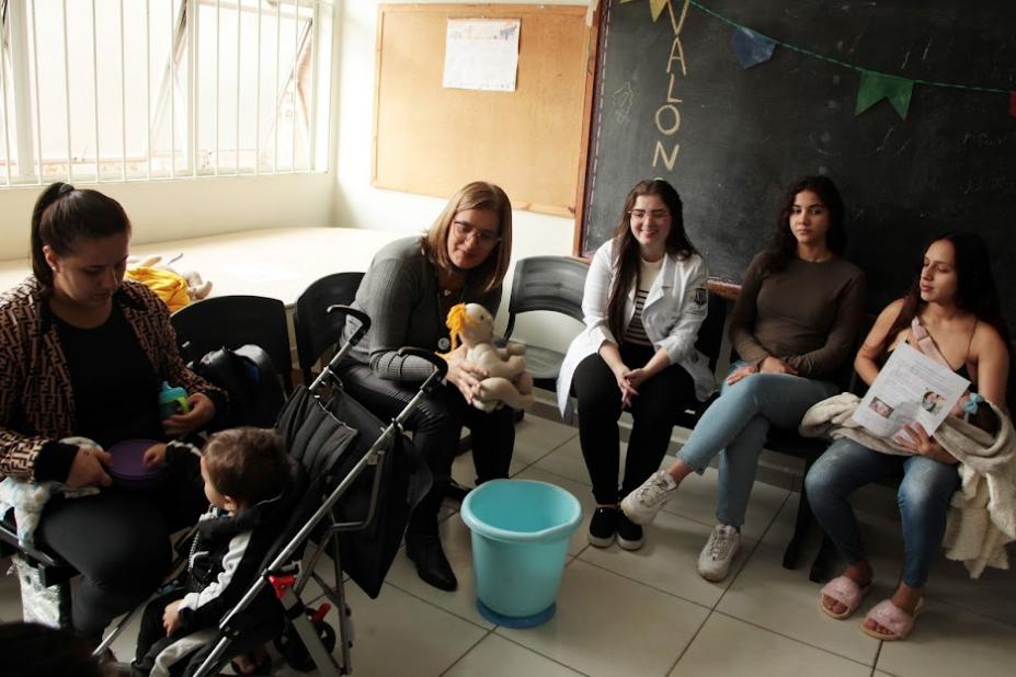 mulher segura boneco próximo a um balde. Mães em torno orbservam. #paratodosverem