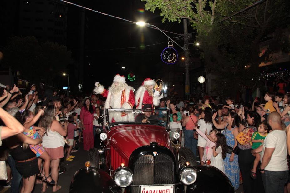 papai e mamãe noel está dentro de calhambeque. Grande público está em torno acenando. #paratodosverem