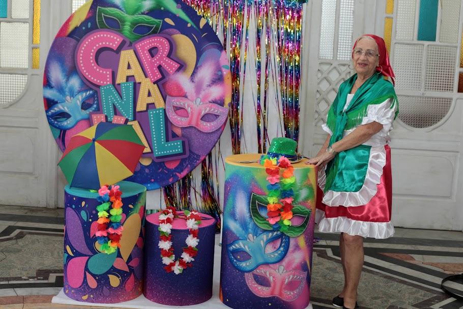 Voluntária está em pé diante de artefatos de carnaval e um banner. Ela está usando uma fantasia que lembra roupas típicas portugues, com avental na cintura e lenço sobre os ombros. #paratodosverem 