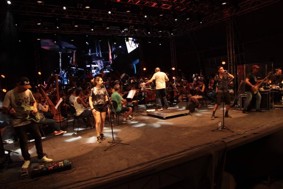 sinfônica está tocando em palco com músicos cantando à frente. #paratodosverem