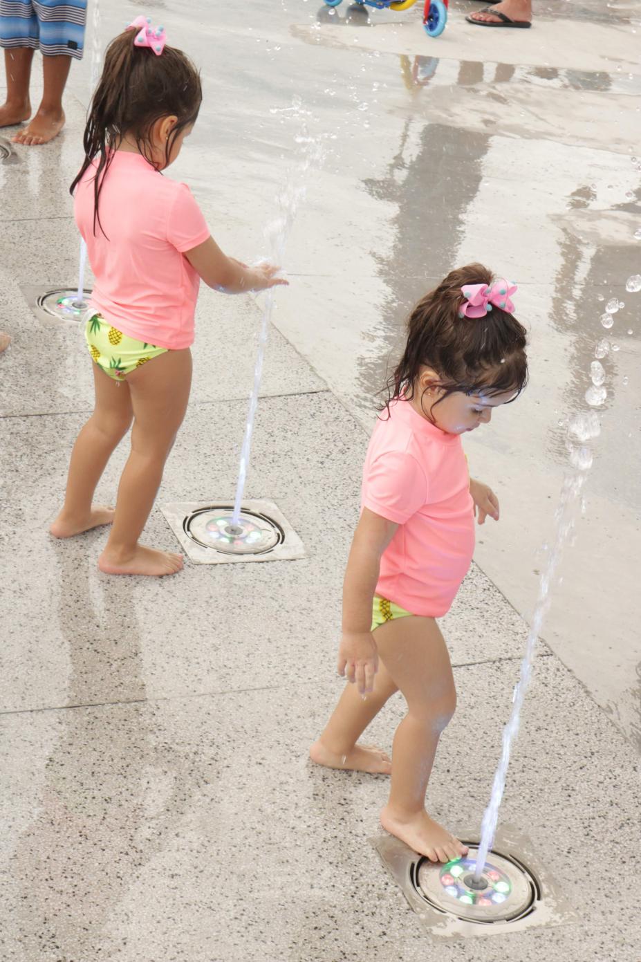 Duas meninas gêmeas estão brincando em dois bicos de fonte interativa. A água está esguichando. #Paratodosverem
