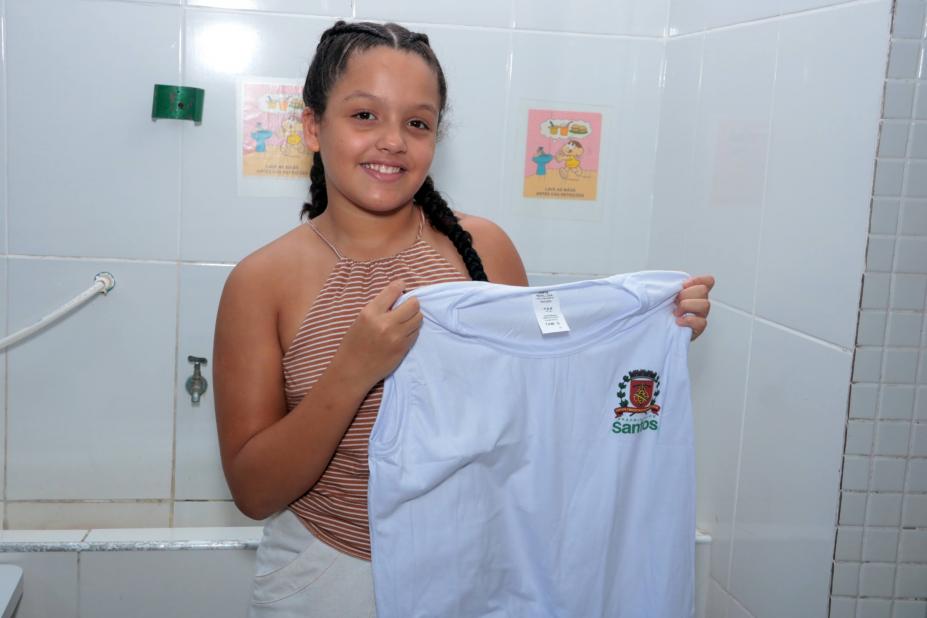 menina sorri para foto segurando camiseta de uniforme aberta pelas duas mãos. #paratodosverem 