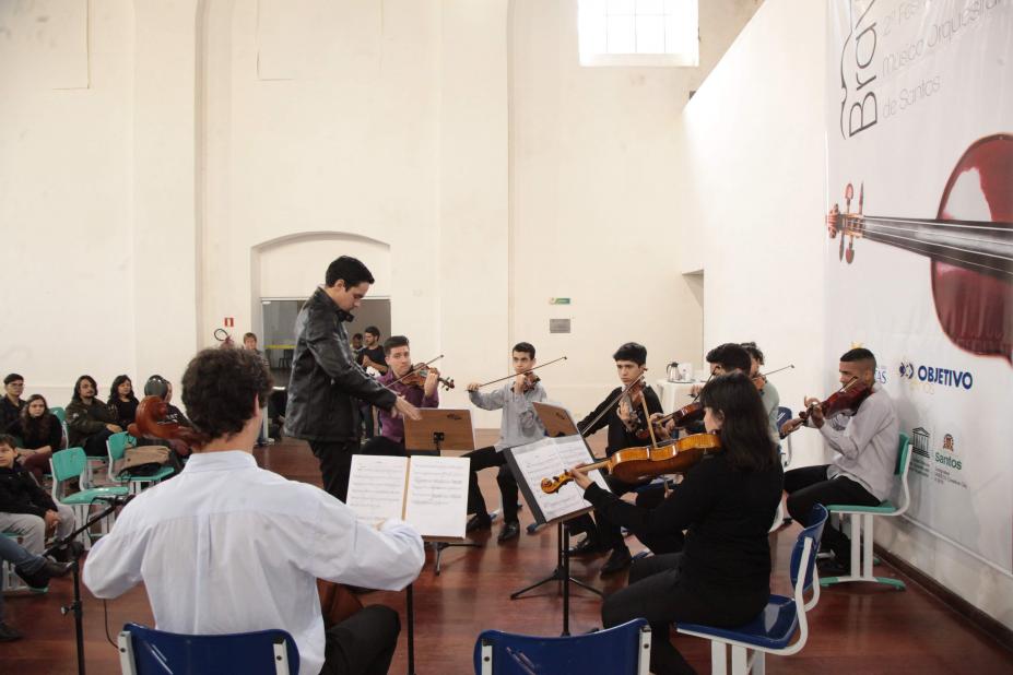 Jovens estão em uma sala, sentados e tocando instrumentos. Alguns têm as estantes à sua frente, com as respectivas partituras. #Pracegover