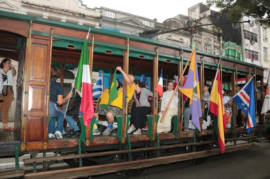 bonde com pessoas carregando bandeiras de vários países por fora do carro. #paratodosverem 