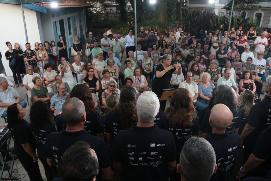 mulher rege coral diante de plateia ao fundo. #paratodosverem