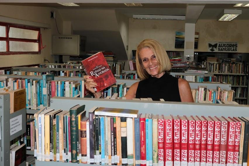 Sueli segura livro entre estantes cheias de livros. ela sorri. #paratodosverem