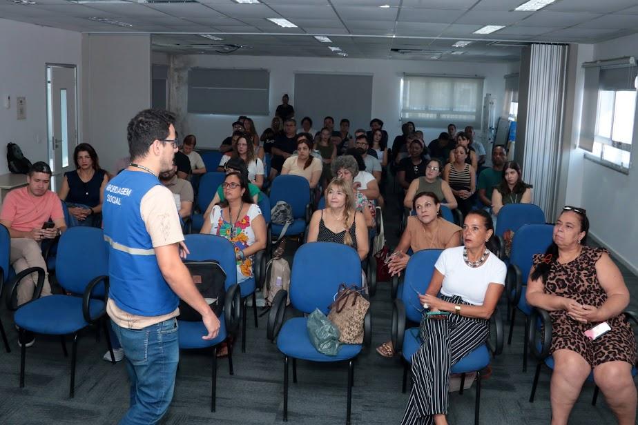 servidores estão sentados em pequeno auditório atentos a um outro servidor que está em pé, de costas para a foto. #paratodosverem