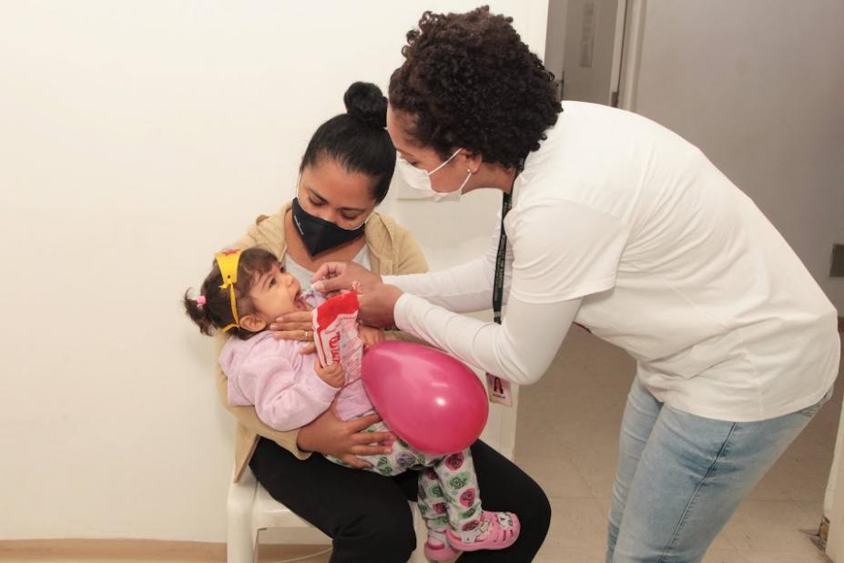 criança está no colo da mãe e mulher dá a gotinha da vacina. #paratodosverem