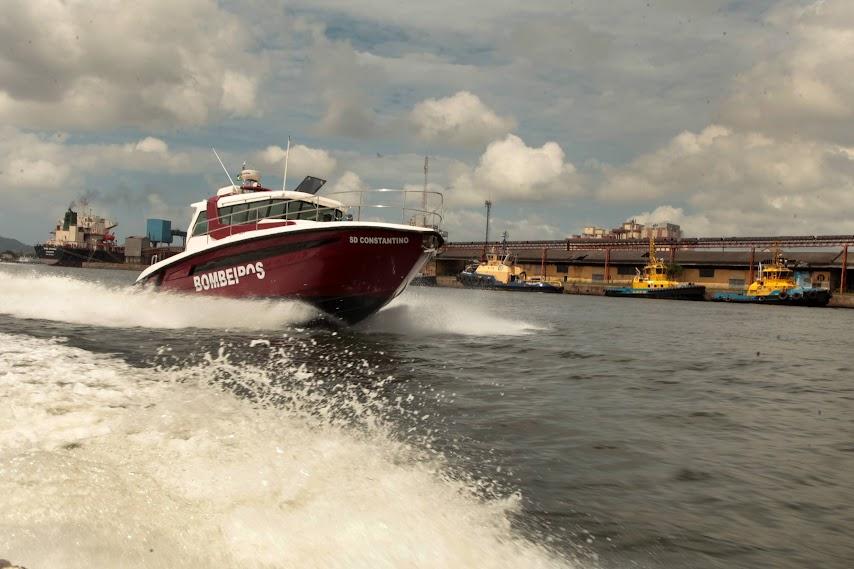 lancha dos bombeiros no mar em movimento com armazéns portuários ao fundo. #paratodosverem