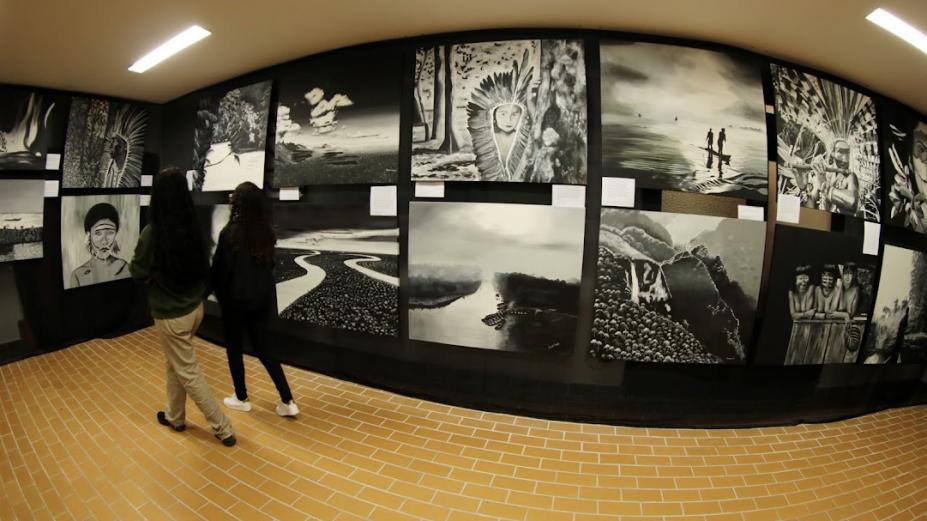 várias pinturas em preto e branco expostas em um painel. Duas pessoas observam a exposição. #paratodosverem 