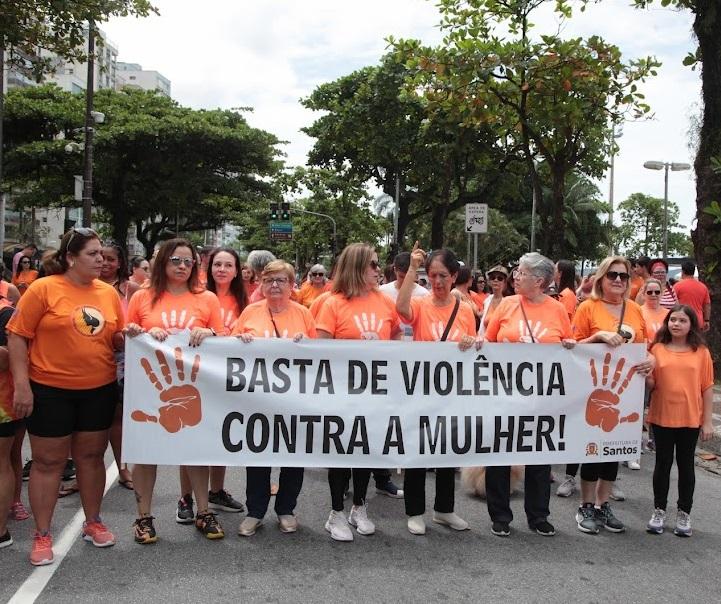 mulheres vestidas com camiseta na cor laranja estão em avenida e as da frente seguram uma faixa onde se lê basta de violência contra a mulher!#paratodosverem