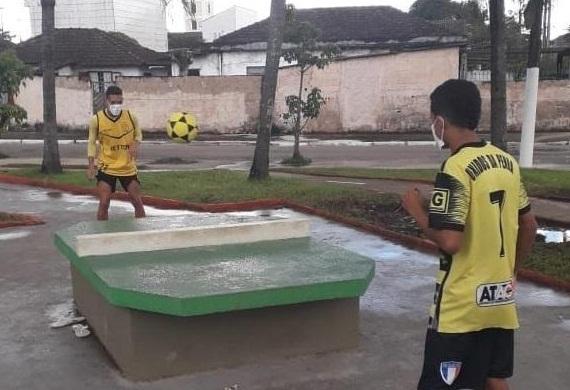 futmesa no centro de uma praça com dois jovens jogando bola, um em cada lado da estrutura. #paratodosverem