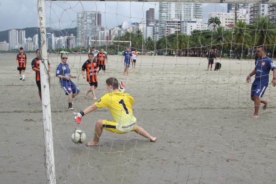 trave de gol em primeiro plano. Atrás,o goleiro tenta defender uma bola. Jogadores estão diante dele olhando a bola entrar na rede. Jogo é na praia. #paratodosverem