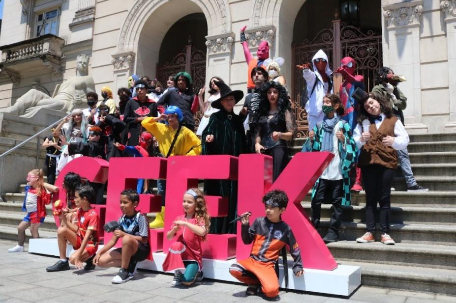 Crianças e jovens fantasiados tiram foto no letreiro Geek na escadaria do paço municipal. #paratodosverem