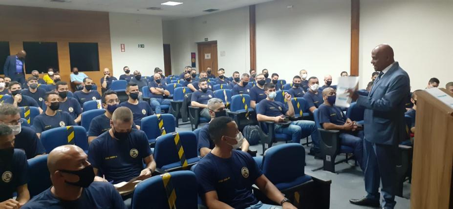 guardas estão sentados em auditório, respeitando o distanciamento, e assistindo palestra de um homem que está em pé. #paratodosverem