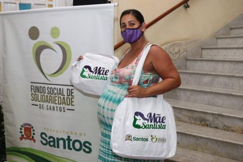 gestante está em pé ao lado de banner do fundo social de solidariedade. Ela segura sacola do programa mãe santista. #paratodosverem