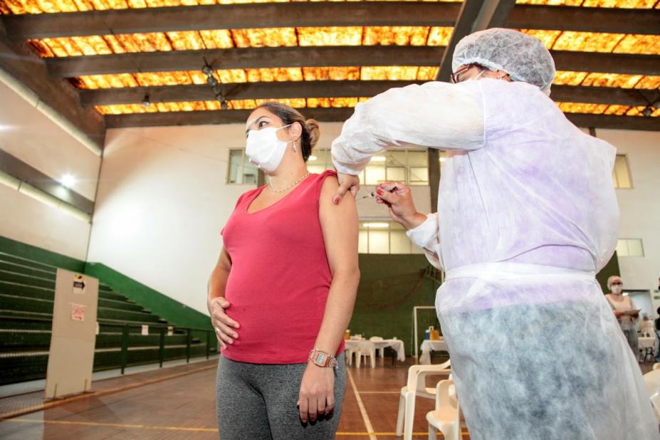 GRAVIDA ESTÁ ACARICIANDO A BARRIGA ENQUANTO É VACINADA. A vacinação ocorre em um ginásio. #paratodosverem