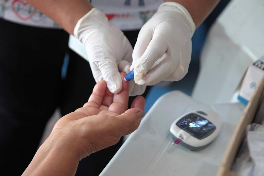 foto em close mostra mãos com luvas tirando gota de sangue da ponta do dedo de outra pessoa. Aparelho de medição de glicemia está ao lado. #paratodosverem