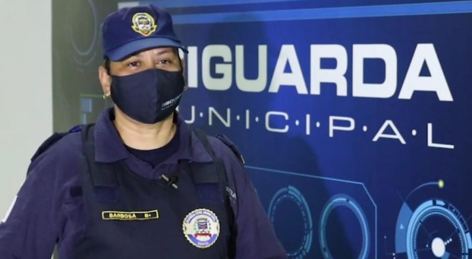 Guarda municipal uniformizada e máscara em frente a painel com o nome da corporação. #pracegover