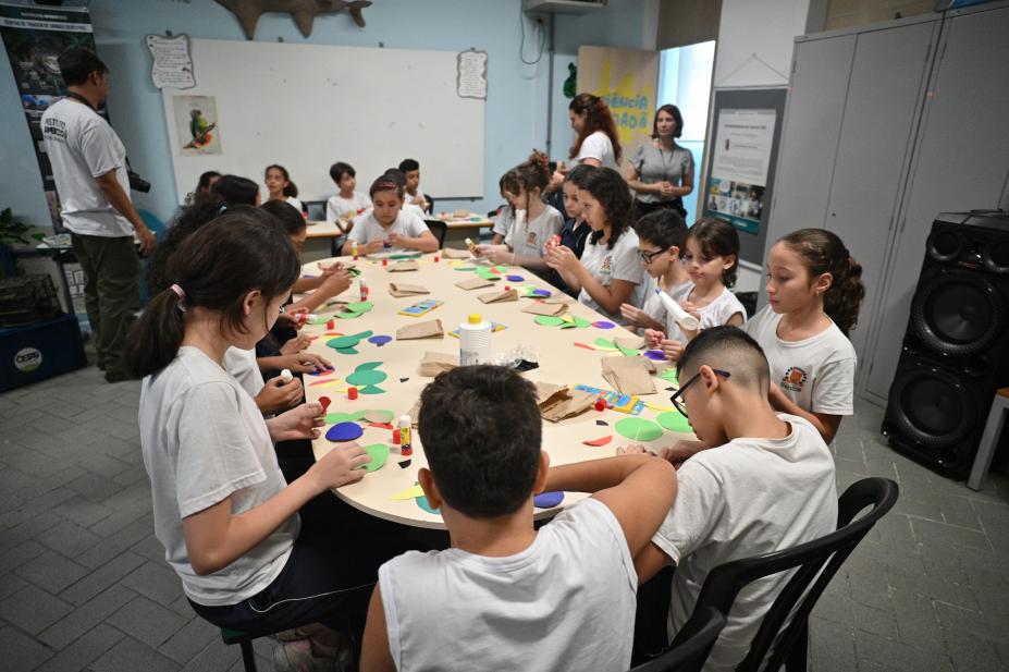 alunos recortam papeis em cima de mesa. #paratodosverem