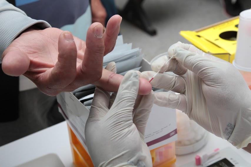 mãos com luvas colhem gota de sangue da ponta do dedo de uma pessoa. Foto em close. #paratodosverem