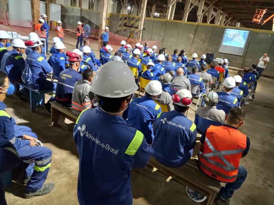 auditório cheio de operários de costas para a foto. Eles estão atentos a um homem que fala ao fundo, ao lado de um telão. Todos os operários usam uniforme e capacete. #paratodosverem