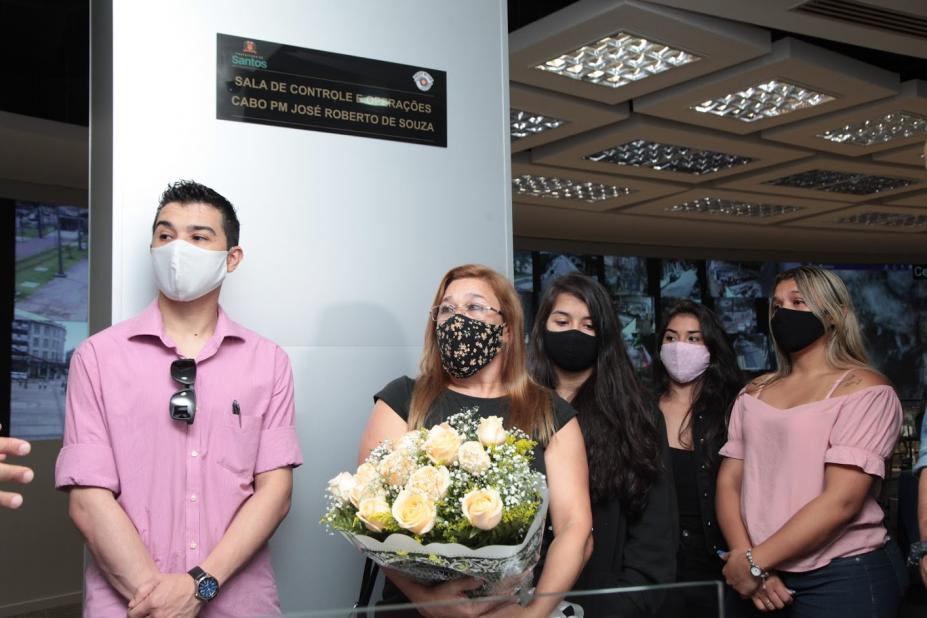 Placa com nome do homenageado afixado na parede. À frente, a viúva segura ramalhete de flores. Os filhos estão ao lado. #Paratodosverem