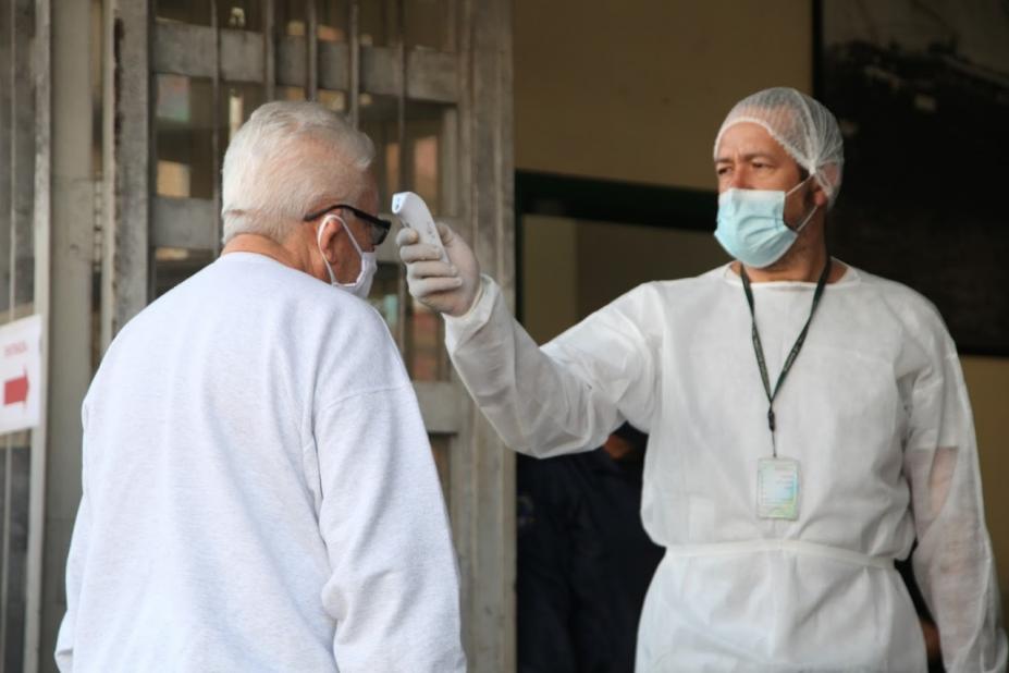 Recepcionista hospitalar, usando máscara, gorro e luva, mede temperatura de idoso com termômetro de testa. #Paratodosverem