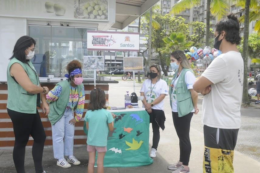 Agentes educativas orienta menina, acompanhada do seu pai, em estande na Fonte do Sapo. #pracegover