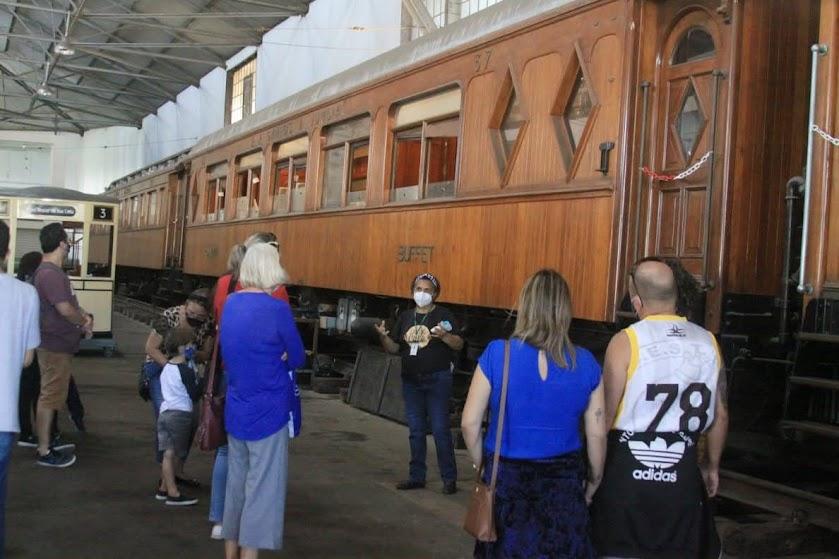 pessoas estão paradas ouvindo orientações de guia diante de antigo carro ferroviário. #paratodosverem 