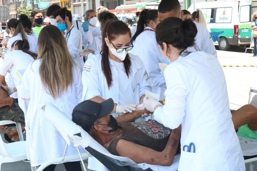 mulheres usando avental branco fazem curativo no braço de homem deitado em maca. #paratodosverem