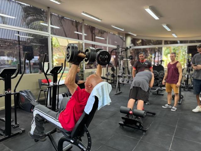 homem está sentado em banco levantando pesos com os dois braços. Ao fundo, um jovem faz exercício num equipamento para lombar. #paratodosverem 