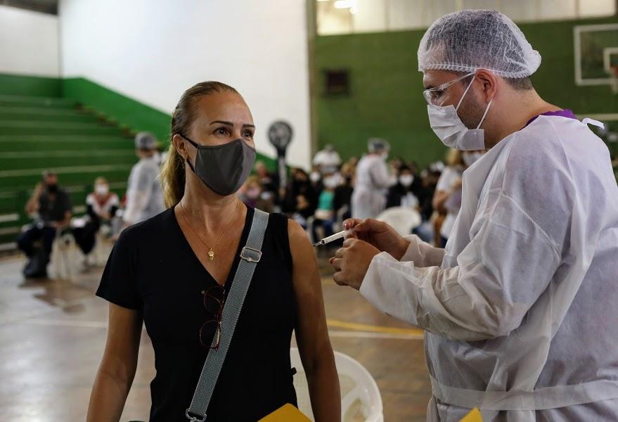Mulher com máscara ao centro recebe aplicação da vacina #paratodosverem