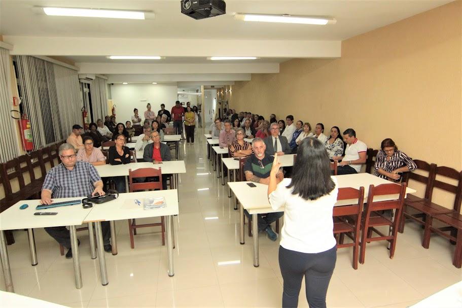 mulher de costas para a foto está falando a pessoas sentadas em uma sala de reuniões. #paratodosverem 