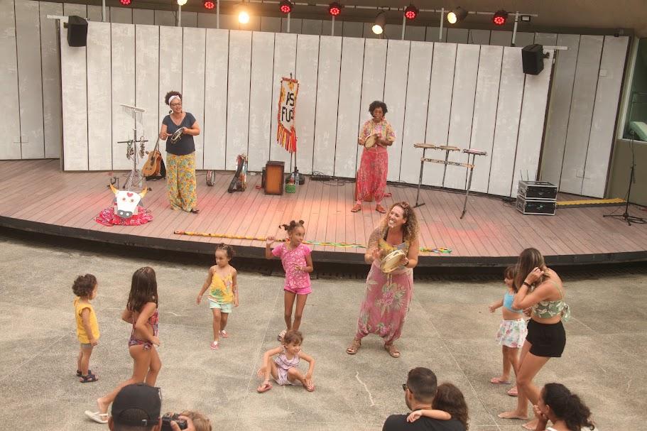 mulheres cantam e tocam em palco ao fundo. À frente, no chão, crianças brincam. #paratodosverem 