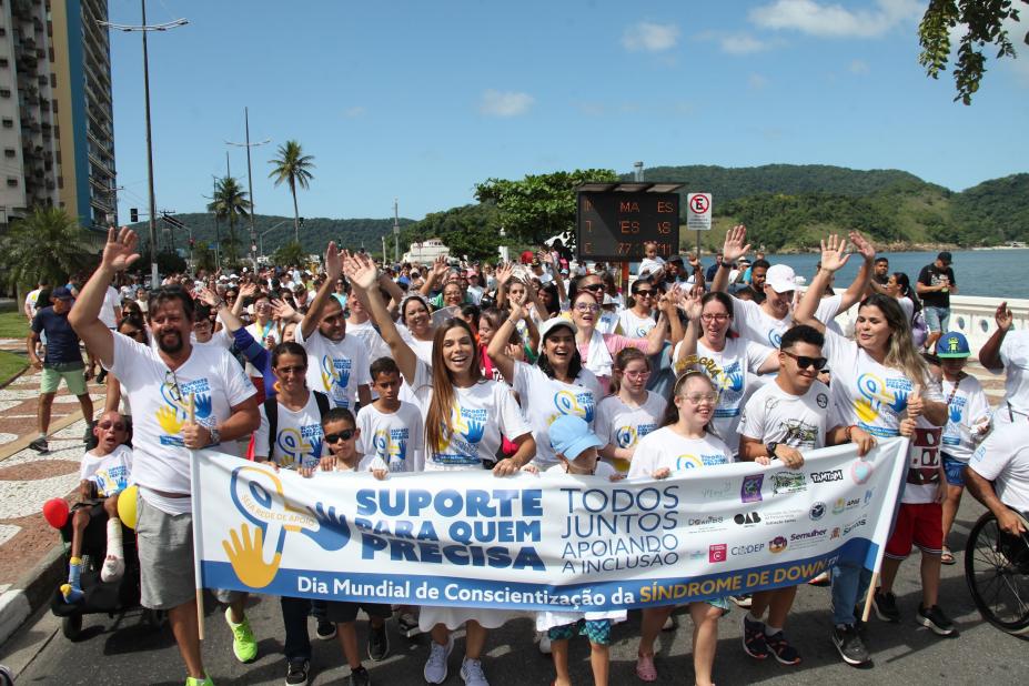 pessoas caminham com camisetas iguais e segurando uma faixa. #paratodosverem 