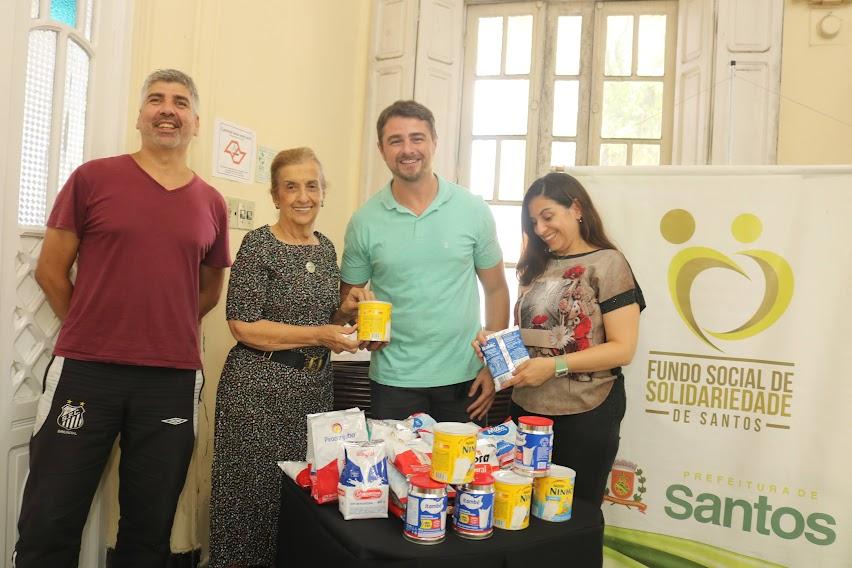 quatro pessoas em pé em torno de uma mesa onde estão latas e pacotes de leite em pó. Ao fundo, um banner do fundo social. #paratodosverem